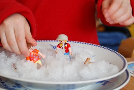 Symbolfoto Mutterschutz. Kinderhände spielen mit Playmobil-Figuren, die in einem Teller stehen, der voller Schnee ist