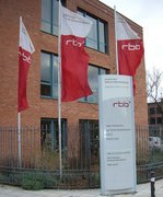 Gezeigt wird ein Eingang des RBB-Studios in Potsdam mit drei RBB-Fahnen, die im Wind flattern und einem Hinweisschild am Eingang.